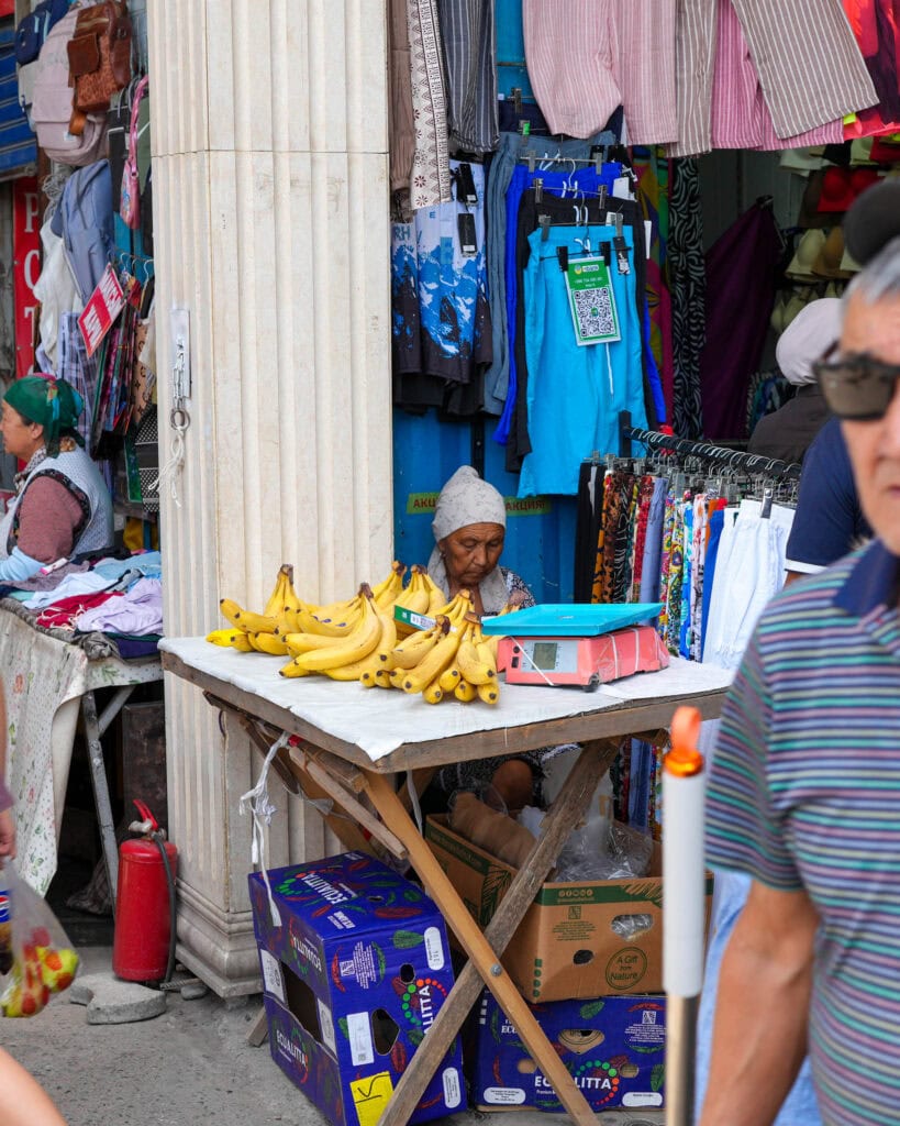 Eine ältere Frau verkauft Bananen an einem Tisch vor einem Bekleidungsgeschäft, während Menschen auf der Straße vorbeigehen.