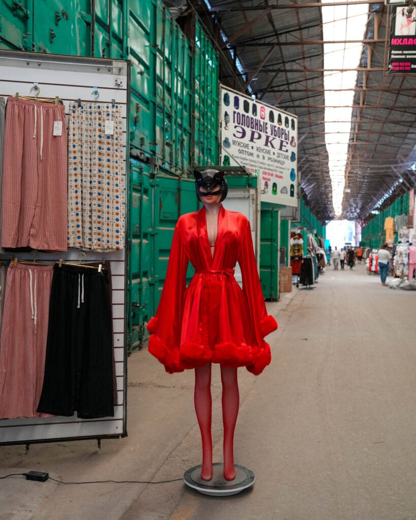 Eine Schaufensterpuppe in einem roten Gewand und einer schwarzen Maske steht in einem Marktgang, daneben hängen Kleidungsstücke zur Ansicht.