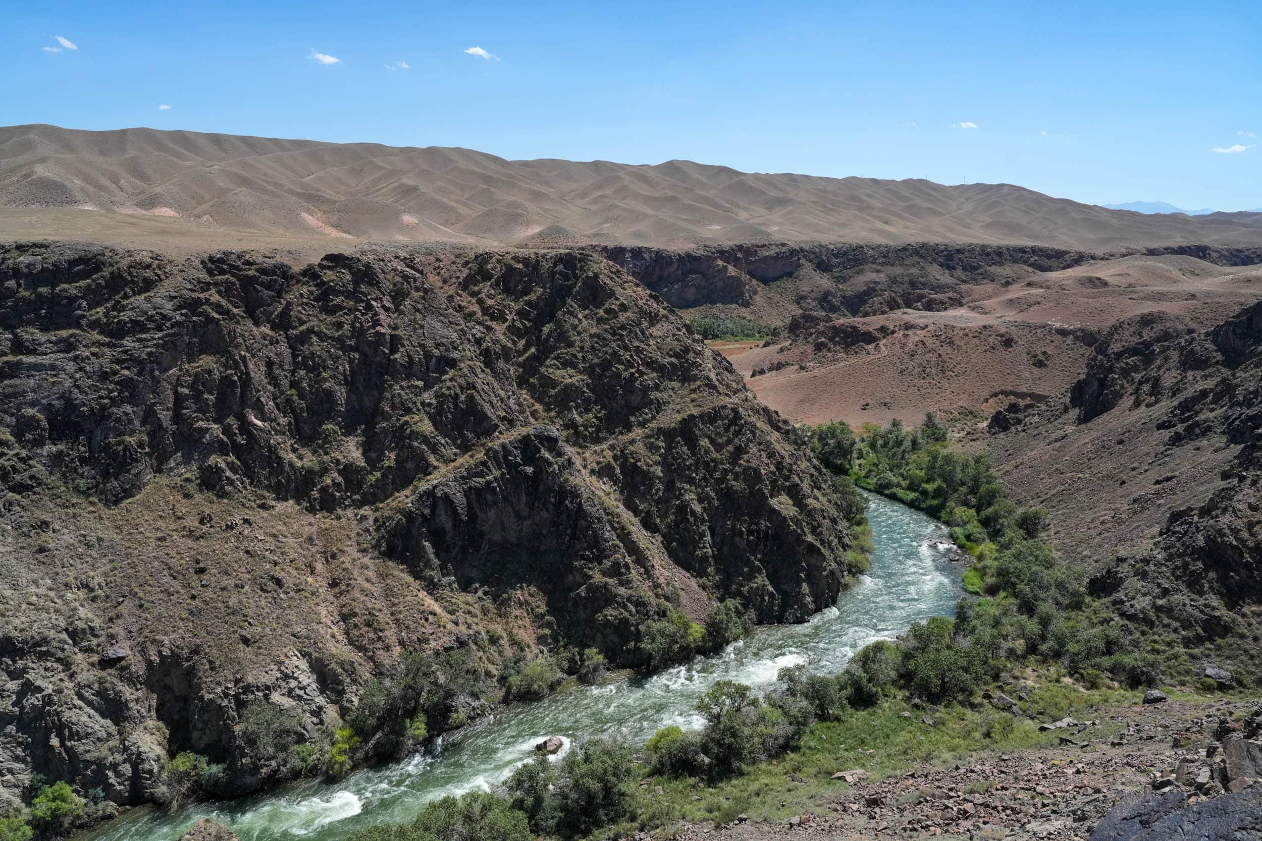 Ein sich schlängelnder Fluss fließt durch eine felsige Schlucht, im Hintergrund erheben sich karge Hügel vor einem strahlend blauen Himmel.