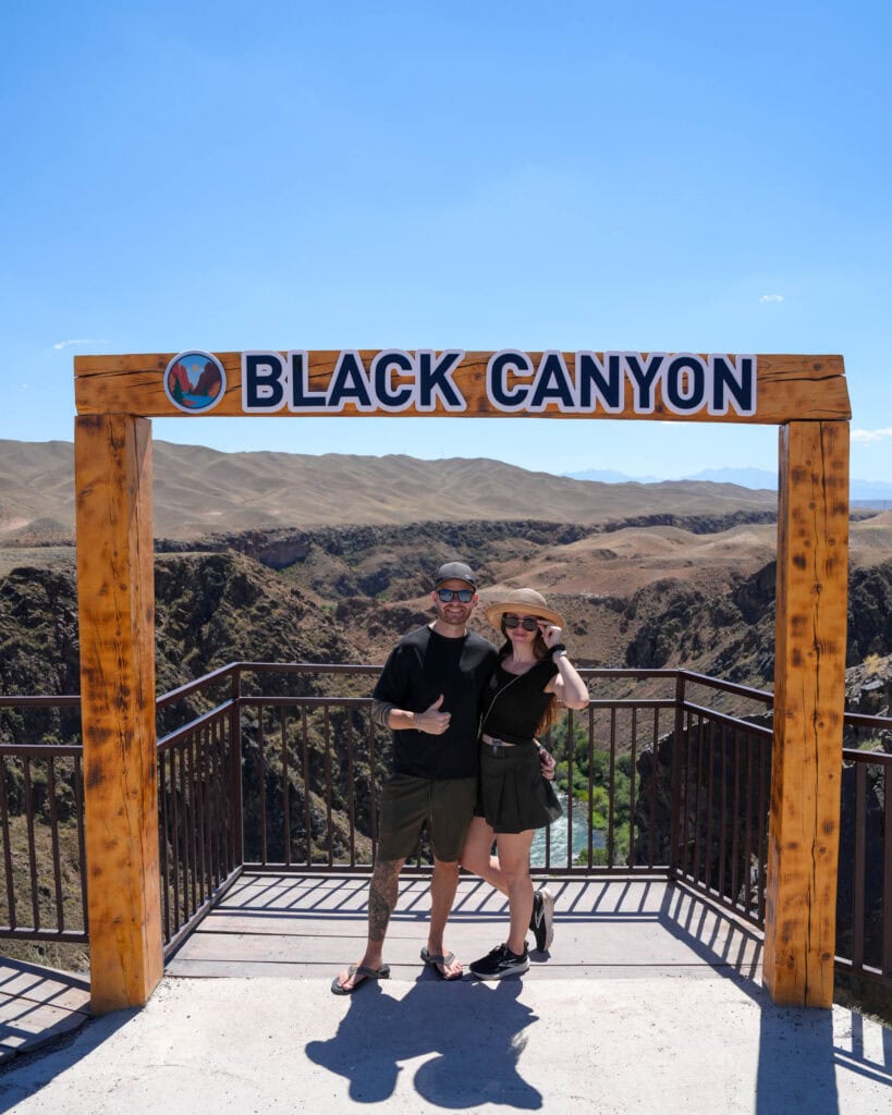 Zwei Personen posieren vor einem „Black Canyon“-Schild, im Hintergrund sind Berge und der blaue Himmel zu sehen.