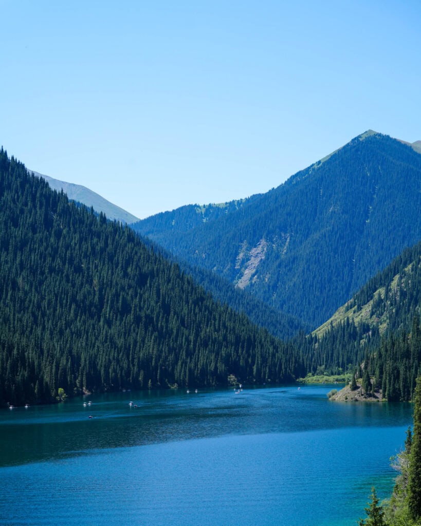 Ein blauer See, umgeben von mit Kiefern bewachsenen Bergen unter einem klaren Himmel, auf dessen Wasser Boote zu sehen sind.