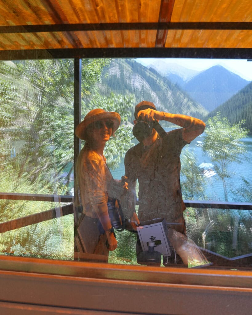 Zwei Menschen spiegeln sich in einem Fenster wider, im Hintergrund sind Bäume, Berge und ein See zu sehen.