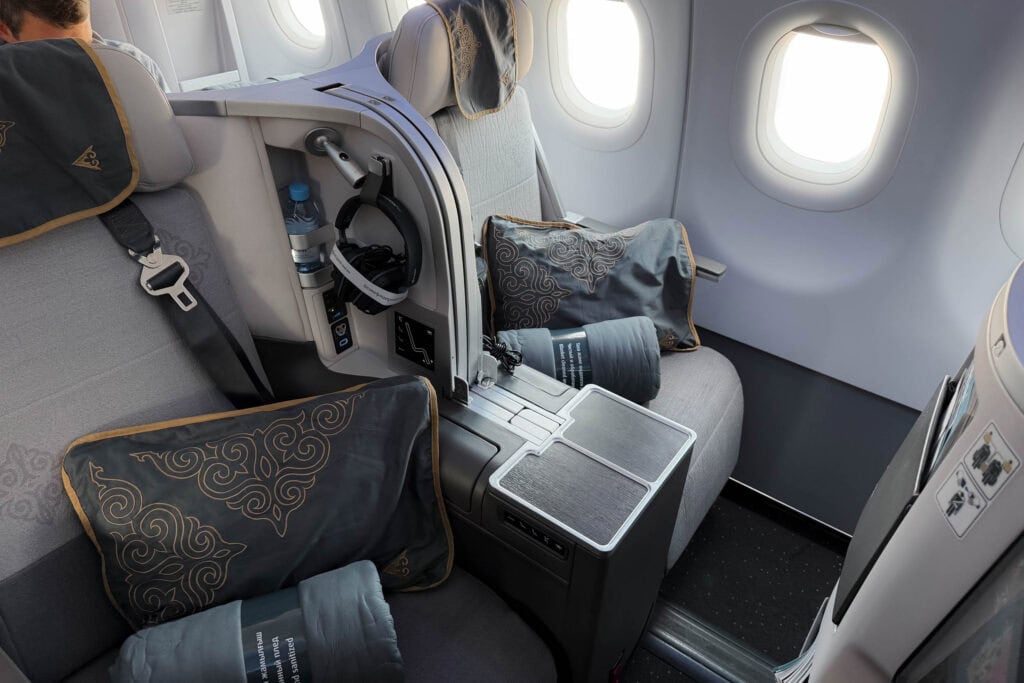 Ein Flugzeugsitz der Business Class mit grauer Polsterung, Kissen, Bettwäsche und großen Fenstern ist zu sehen.