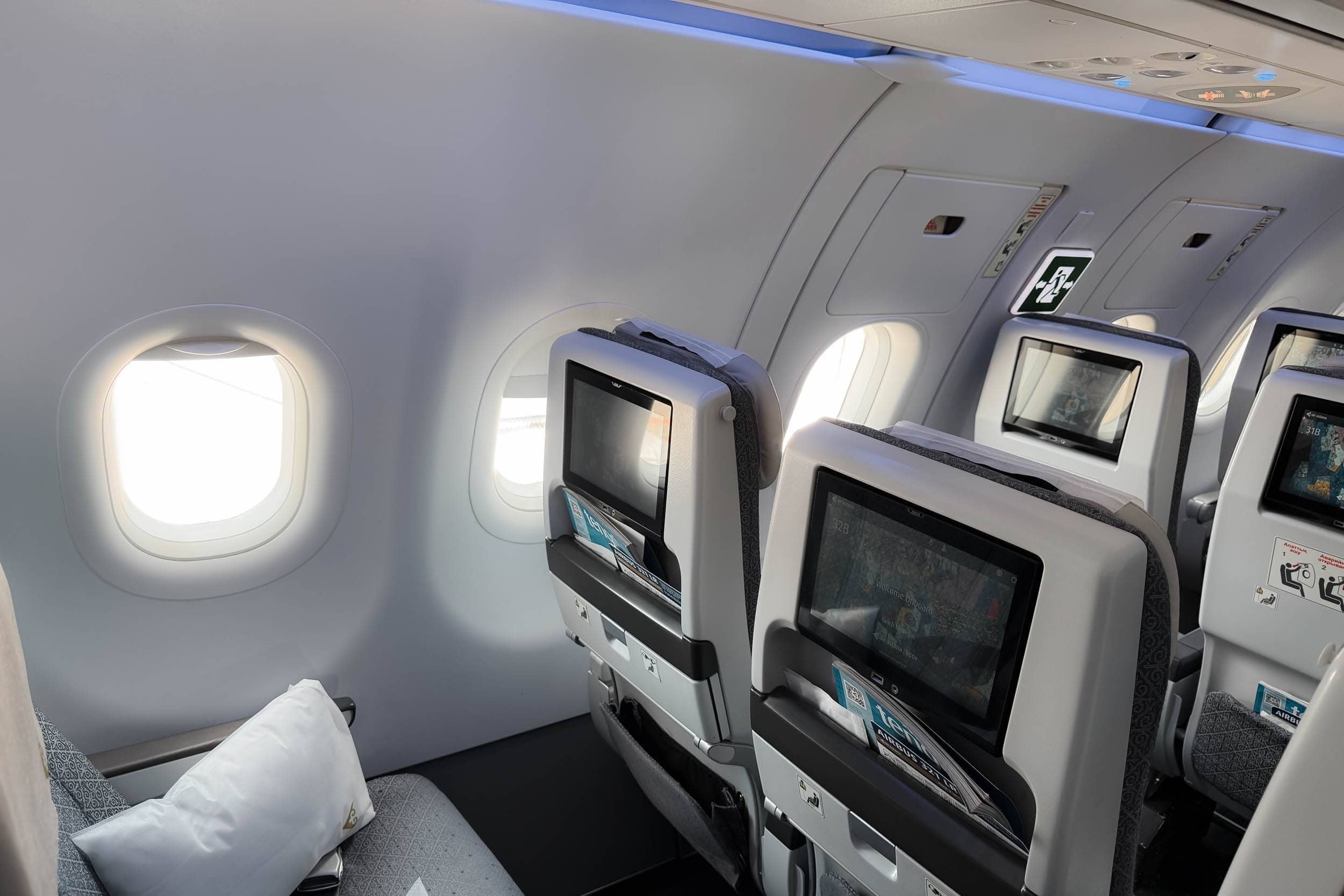 Drei Flugzeugsitze mit Bildschirmen in den Rückenlehnen, hochgeklappten Klapptischen und Kissen, neben zwei Fenstern bei Tageslicht.