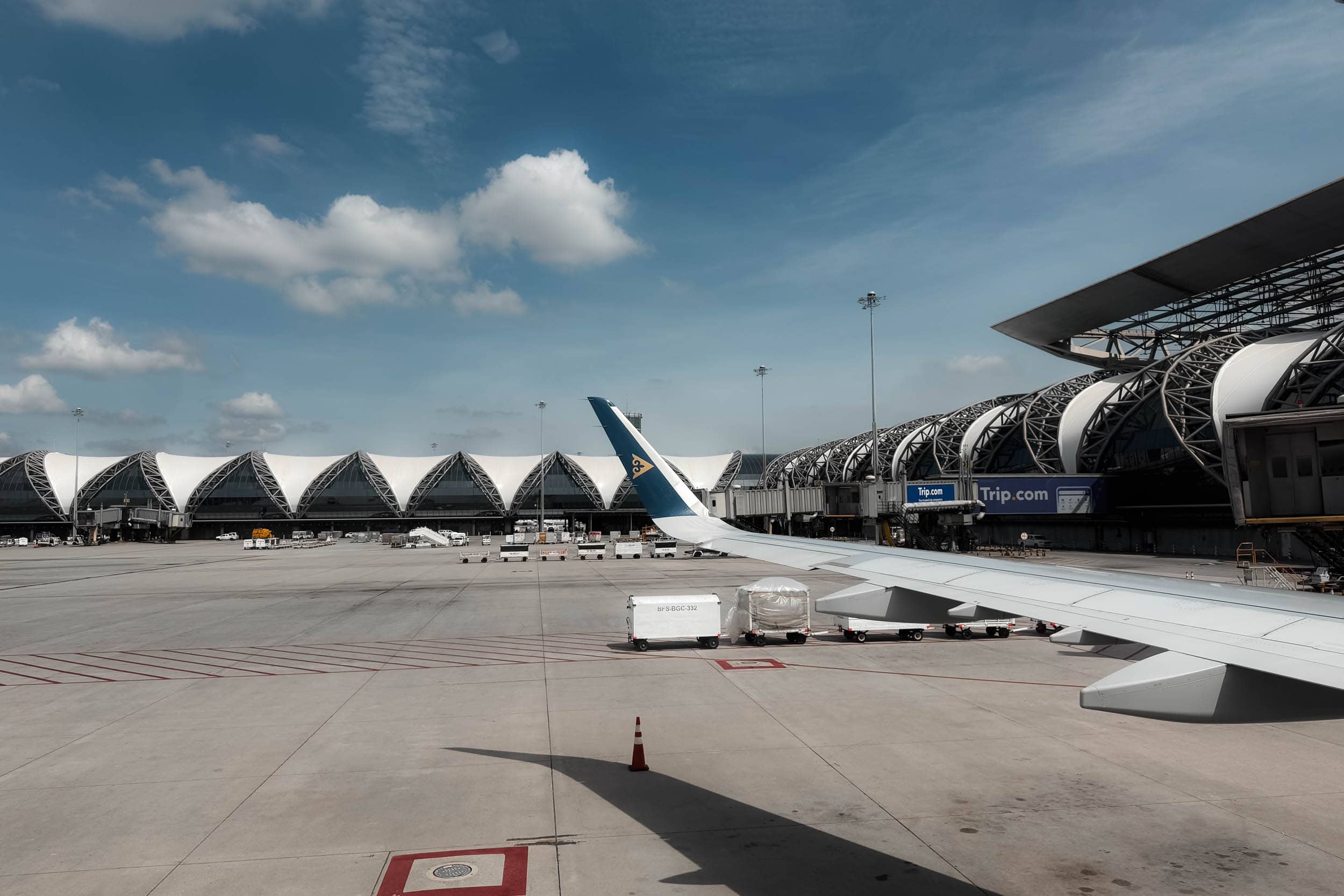 Blick aus dem Flugzeugfenster auf das Flughafenterminal, einen Flugzeugflügel, Frachtwagen und den blauen Himmel mit Wolken.