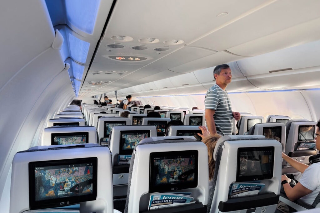 Passagiere, die in der Flugzeugkabine sitzen und stehen, während die Bildschirme in den Rückenlehnen eingeschaltet sind.