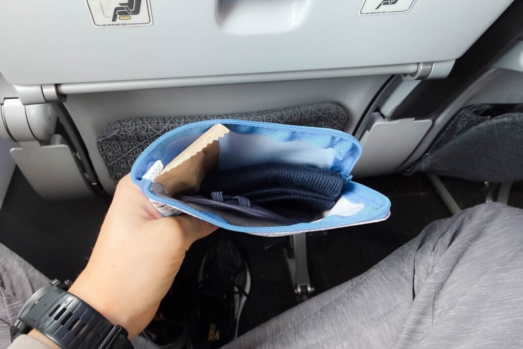 In einem Flugzeug hält eine Hand einen offenen blauen Reisebeutel in der Hand, in dem Socken und eine Schlafmaske zu sehen sind.