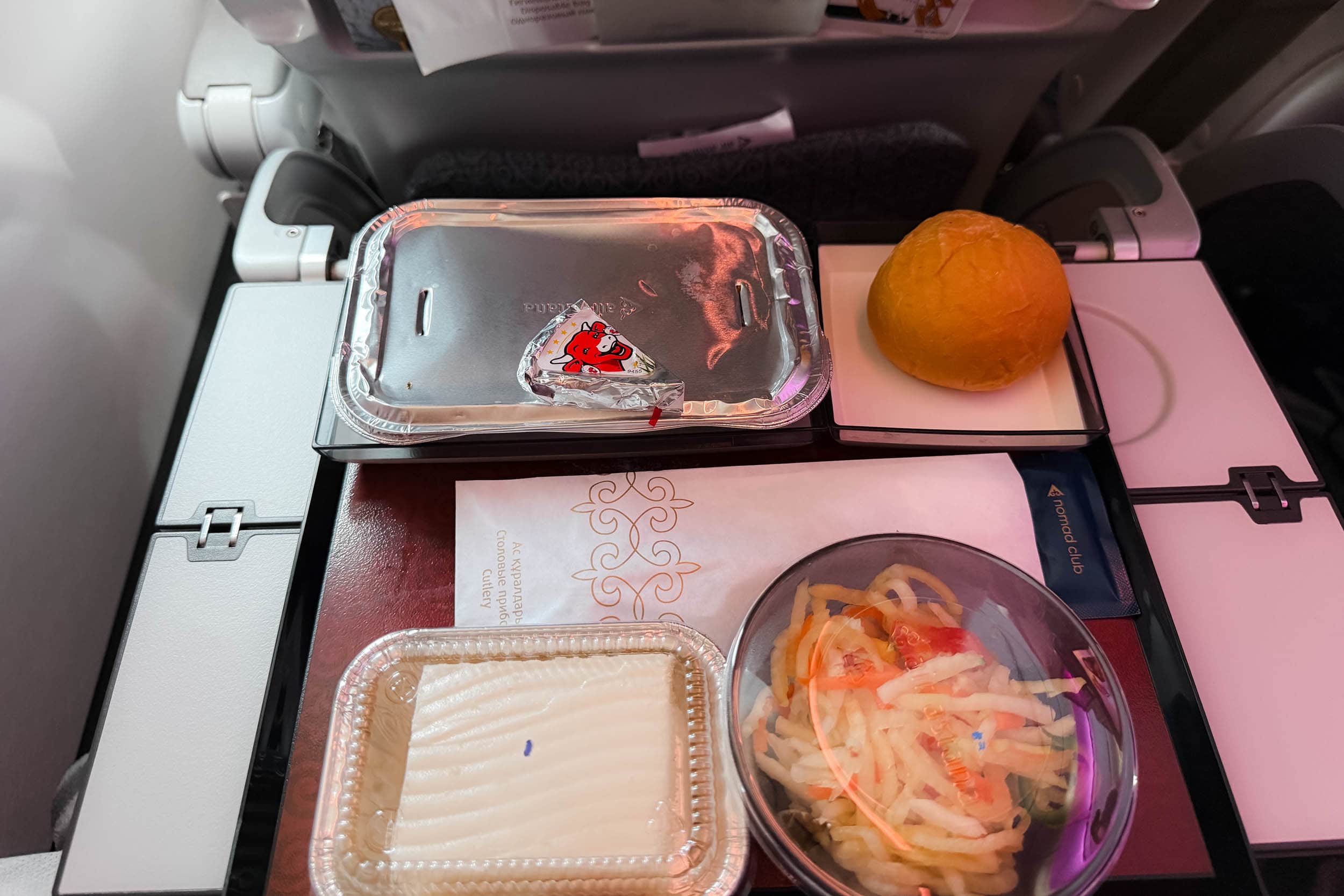 Flugzeugmahlzeit auf einem Tablett mit einem Brötchen, einem abgedeckten Hauptgericht, Salat, Dessert und einer Serviette auf dem Klapptisch eines Flugzeugsitzes.