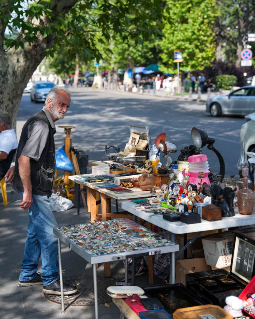 An einem sonnigen Tag stöbert ein Mann an einem Stand auf einem Flohmarkt im Freien, der mit verschiedenen Vintage-Artikeln und bunten Gegenständen gefüllt ist.