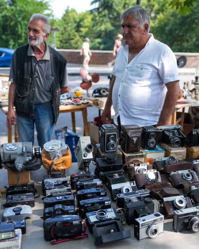 An einem sonnigen Tag stehen zwei Männer auf einem Markt im Freien hinter einem Tisch, auf dem zahlreiche Oldtimer-Kameras ausgestellt sind.