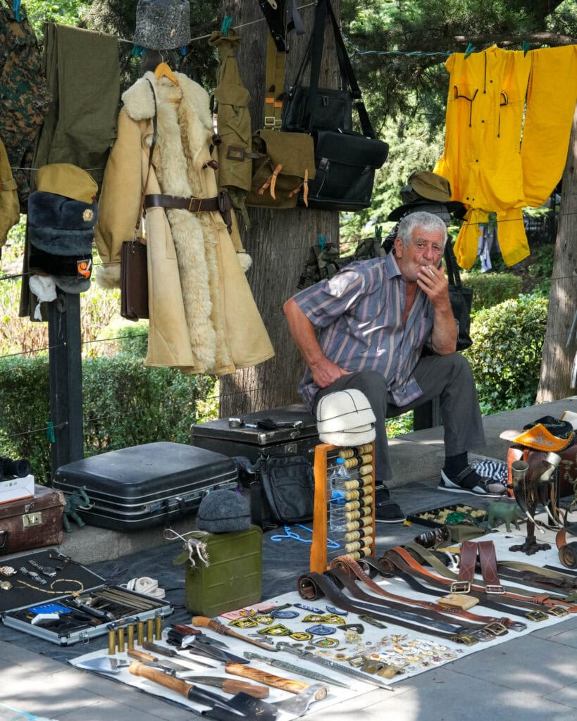 Ein älterer Mann sitzt an einem Stand auf einem Markt im Freien und verkauft Vintage-Kleidung, Taschen, Gürtel und Militaria.