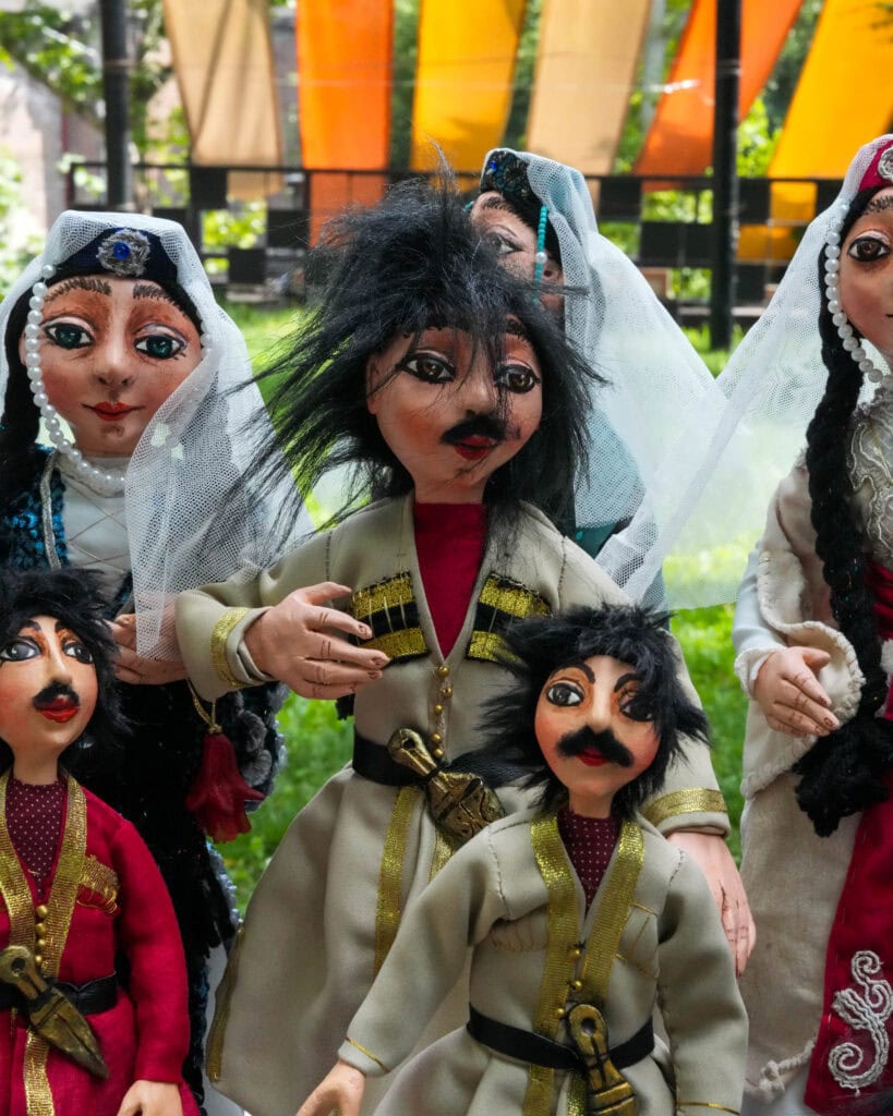 Handgefertigte Puppen in traditioneller Kleidung mit bemalten Gesichtern, im Freien vor einem Hintergrund aus bunten Stoffen.