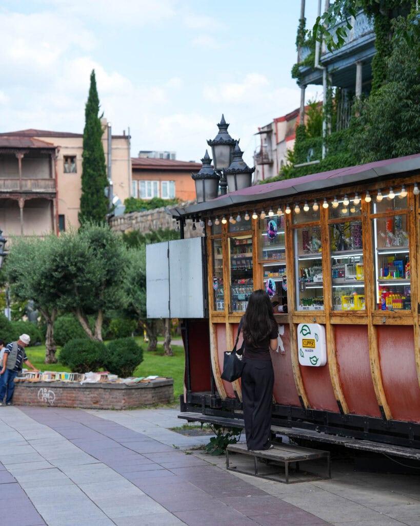 Eine Frau kauft an einem Kiosk ein, der aus einer alten Straßenbahn umgebaut wurde und auf einem Stadtplatz steht, in der Nähe von Grünflächen und Gebäuden.