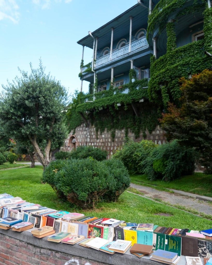 Bücher, die an einer Backsteinmauer vor einem großen, mit Efeu bewachsenen Haus mit Garten und Bäumen ausgestellt sind.