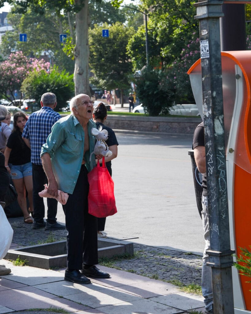 Ein älterer Mann mit einer roten Tasche und einem Stofftier schreit in der Nähe eines öffentlichen Telefons auf einer sonnigen Straße.