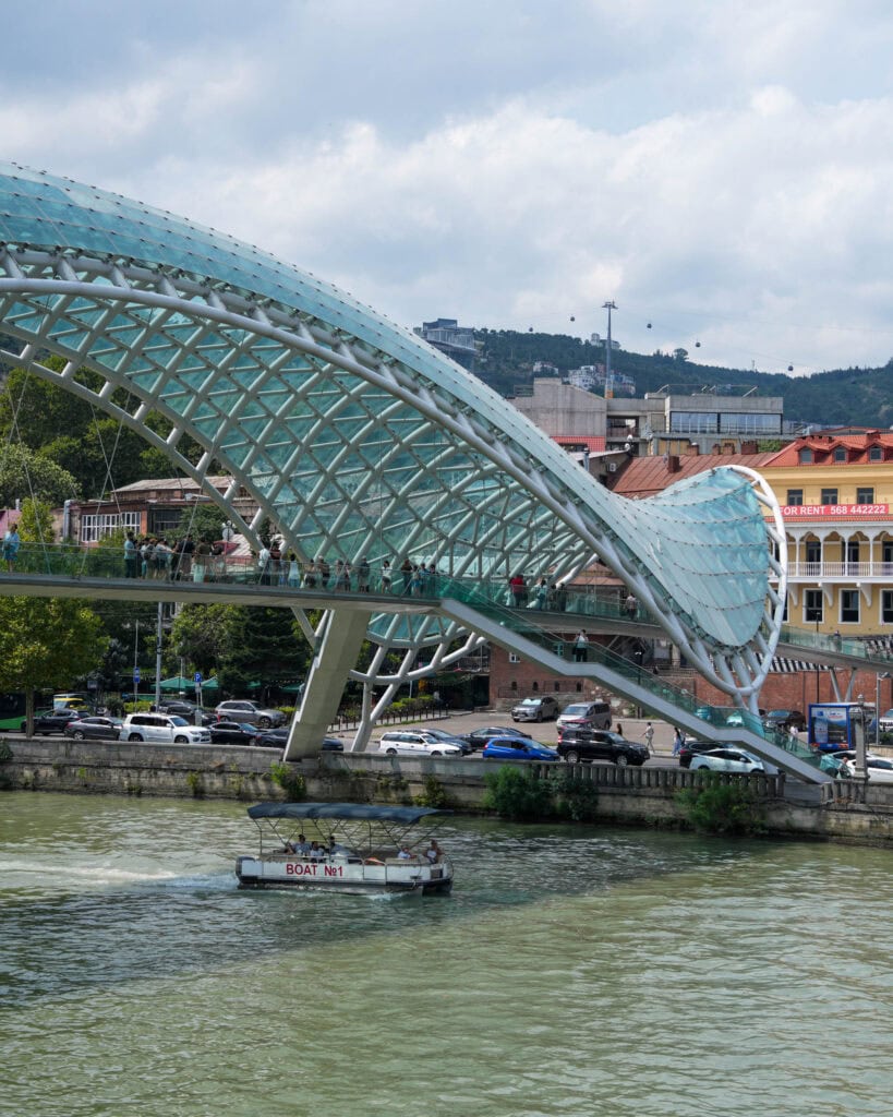 Moderne Glasbrücke über einen Fluss, auf der Menschen spazieren gehen; darunter fährt ein kleines Boot vorbei; im Hintergrund sind Stadtgebäude zu sehen.