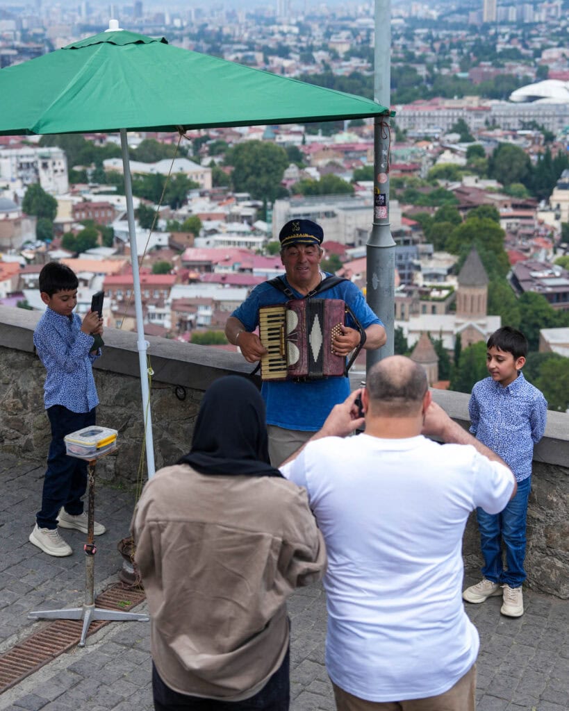 Ein Mann spielt auf einer Aussichtsplattform in der Stadt Akkordeon, während die Leute Fotos machen; zwei Jungen stehen daneben.
