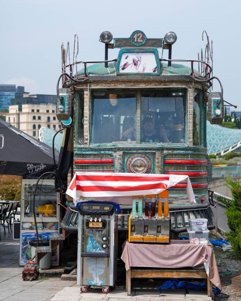Eine alte Straßenbahn, die zu einem Café umgebaut wurde, mit Getränkeautomaten und einem gestreiften Vordach vor dem Eingang sowie Sitzgelegenheiten im Freien.