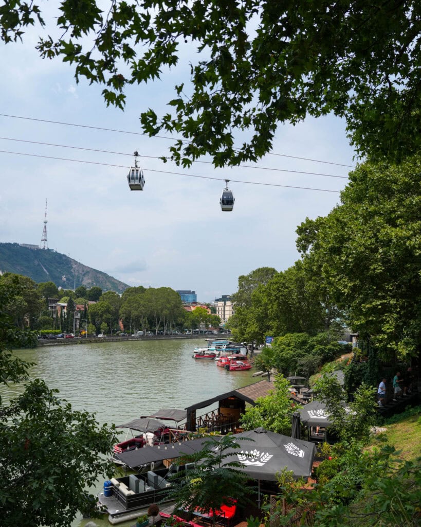An einem bewölkten Tag fahren Seilbahnen über ein Café am Flussufer, Boote und üppige Bäume hinweg, während im Hintergrund Hügel zu sehen sind.