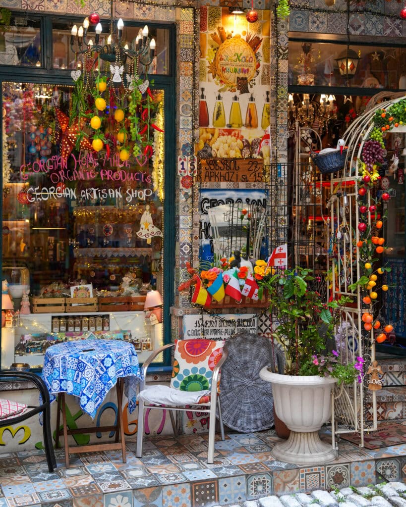 Farbenfrohe Ladenfront eines Kunsthandwerksgeschäfts mit hängenden Dekorationen, Fahnen, bunten Stühlen, Mosaikfliesen und Blumen in Töpfen.