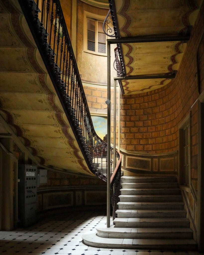 Eine geschwungene Treppe mit kunstvoll verziertem Geländer, Fliesenboden und gestrichenen Wänden in einem warm beleuchteten, historischen Gebäude.
