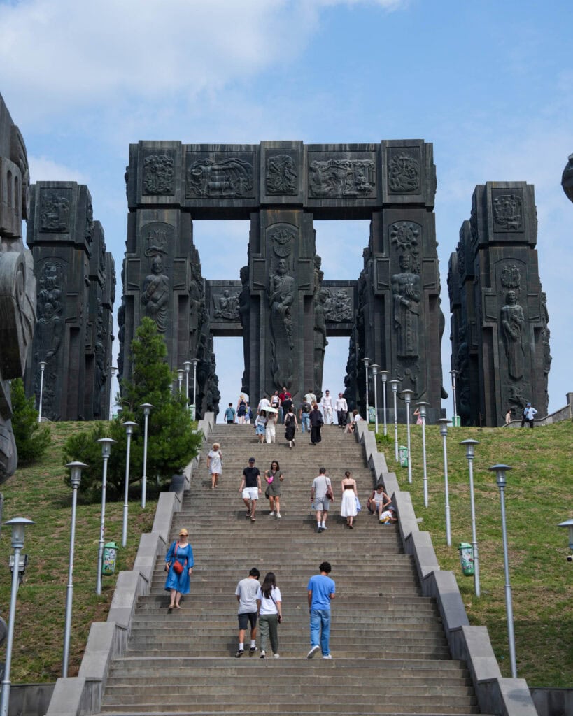 Die Menschen gehen eine breite Treppe hinauf zu einem großen, dunklen Steinmonument mit hohen Säulen und Skulpturen.
