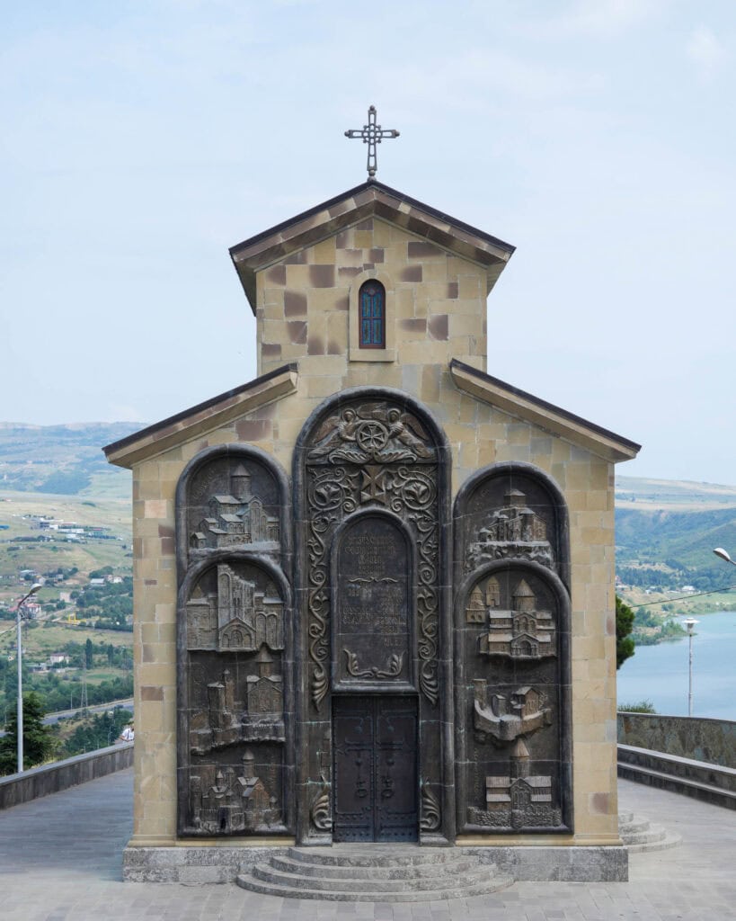 Kleine Steinkapelle mit kunstvollen Metallreliefs an der Fassade, umgeben von einer malerischen Landschaft und einem See.