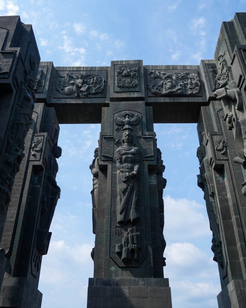 Ein hohes Steinmonument mit detailreichen Mensch- und Tierreliefs vor einem blauen Himmel mit vereinzelten Wolken.