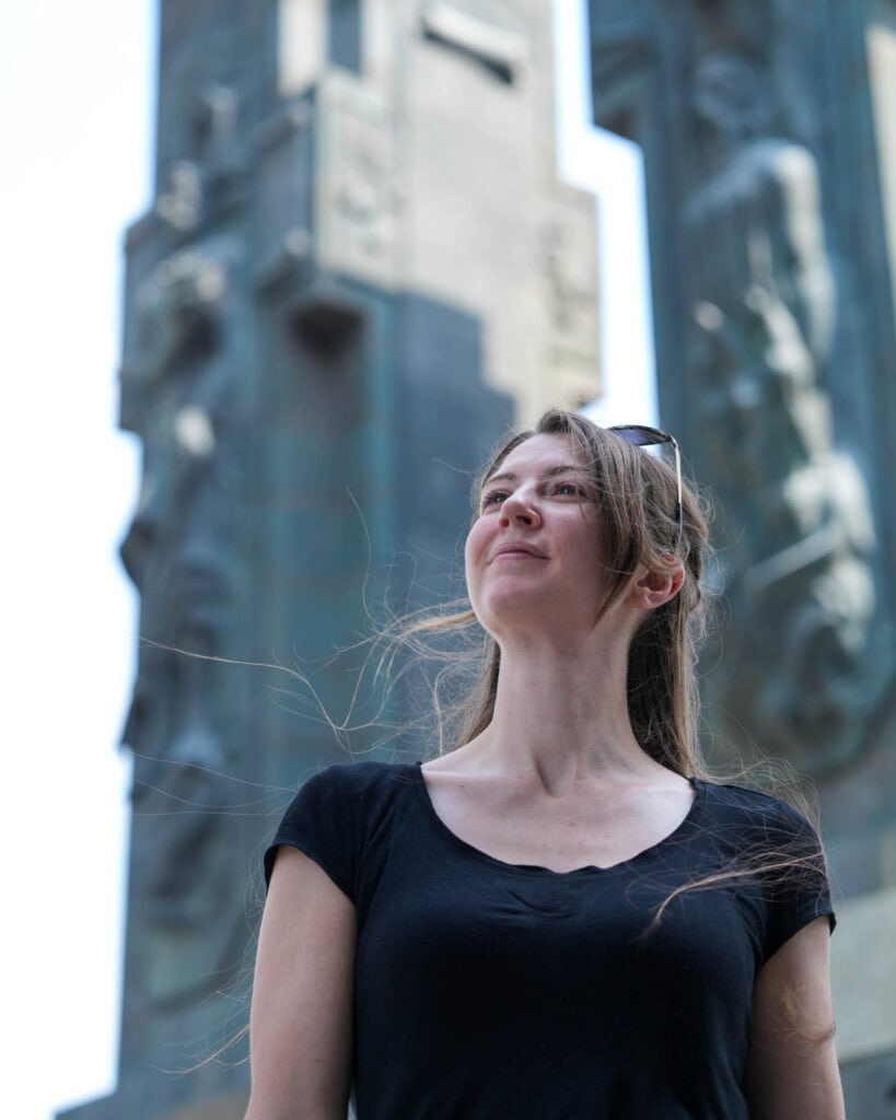 Eine Frau in einem schwarzen Shirt steht draußen und blickt nach oben, im Hintergrund sind hohe Steinskulpturen zu sehen.
