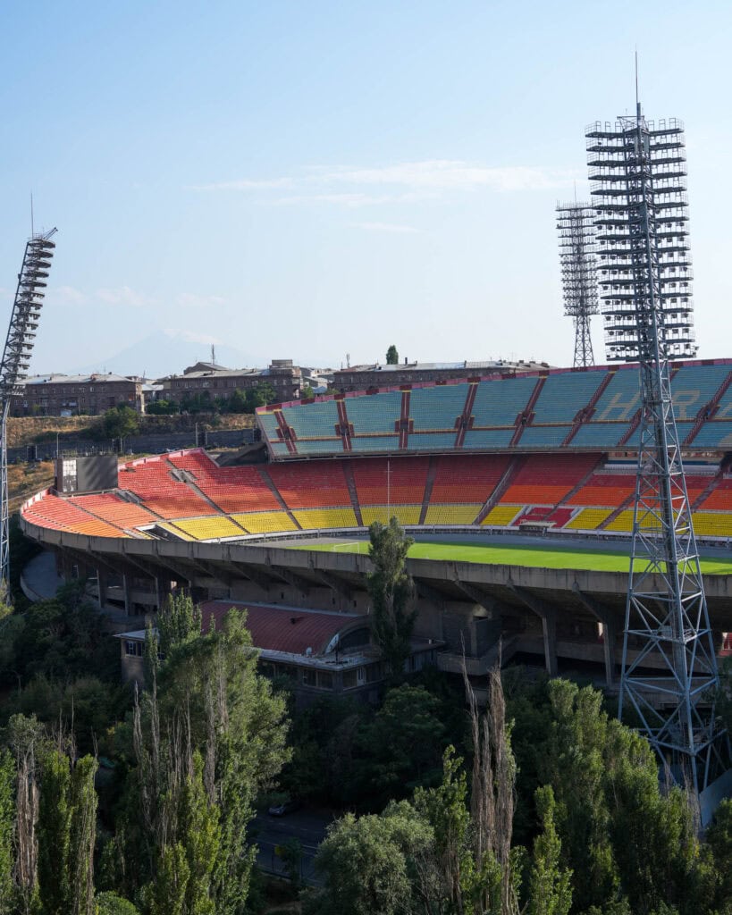 Ein leeres Stadion mit bunten Sitzplätzen, hohen Flutlichtmasten und Bäumen im Vordergrund unter einem klaren Himmel.