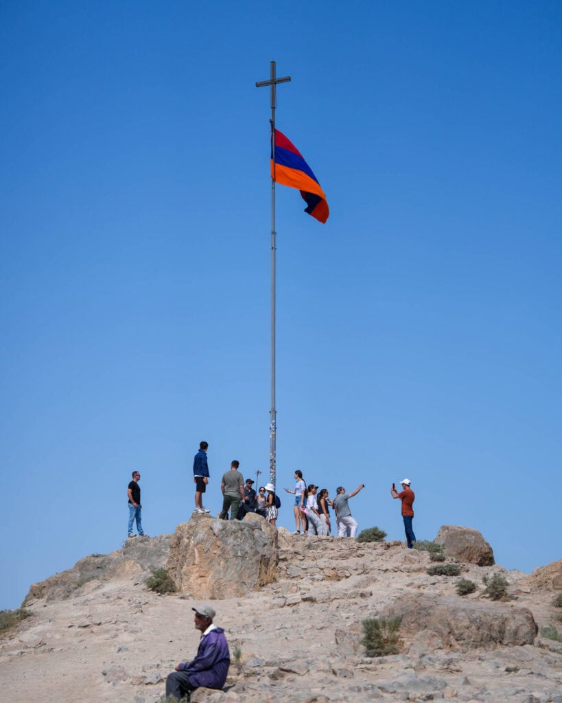 Auf einem Hügel versammeln sich Menschen um eine armenische Flagge und ein Kreuz, während im Vordergrund eine Person etwas abseits sitzt.