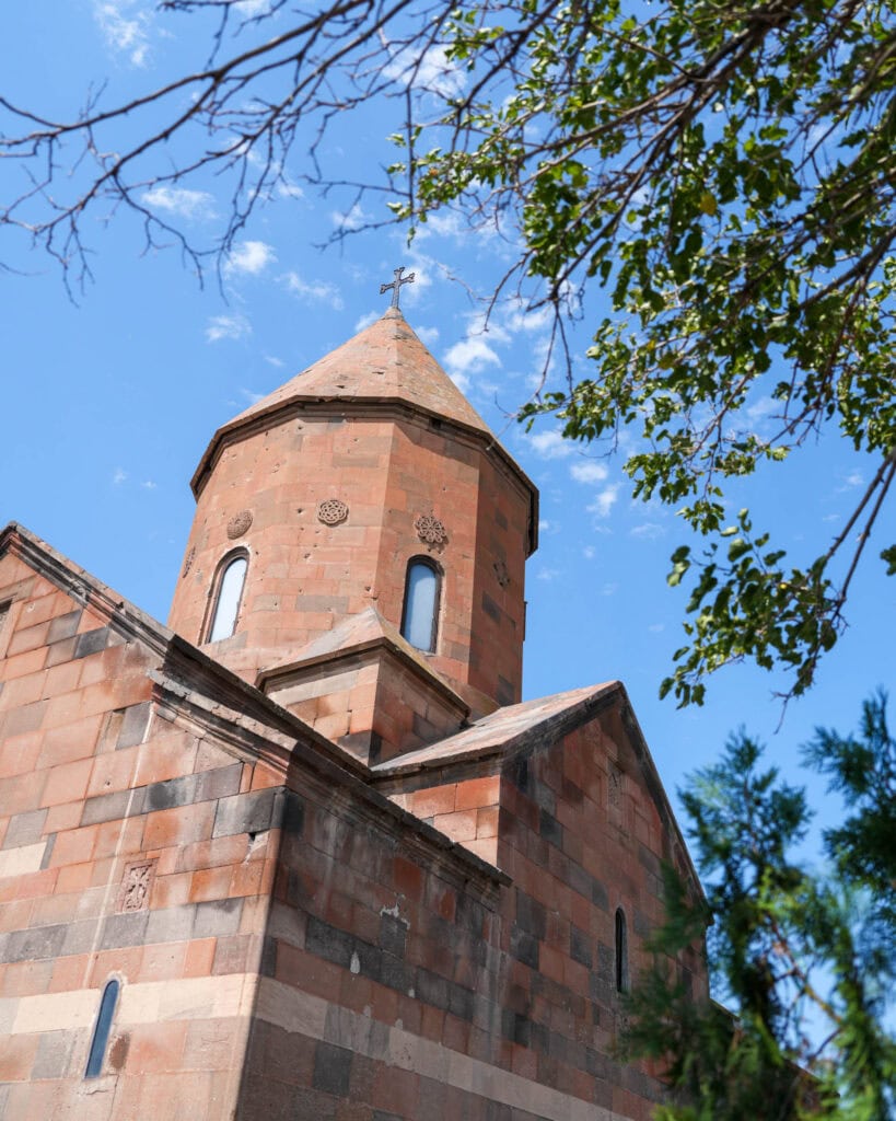 Eine Steinkirche mit einem hohen, spitzen Turm, von unten gesehen, darüber blauer Himmel und Baumzweige.