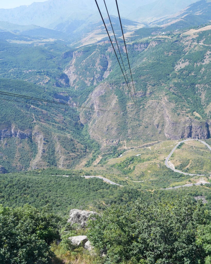 Die Gleise einer Seilbahn führen hinab durch eine üppige Berglandschaft, unterhalb derer sich kurvenreiche Straßen und grüne Täler erstrecken.