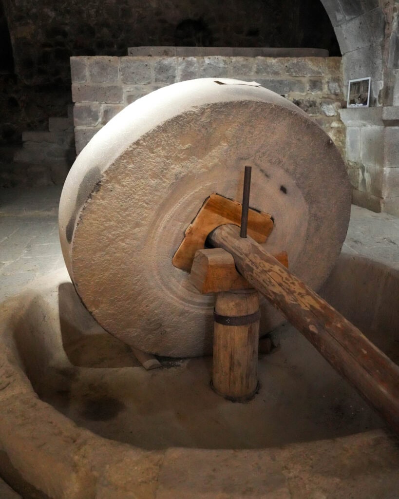 Großes Mühlrad aus Stein mit Holzgriff, eingebettet in einen Steinraum mit gewölbten Wänden und gedämpfter Beleuchtung.