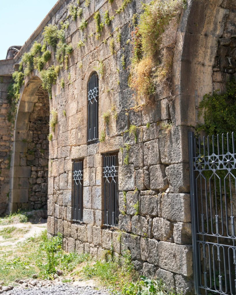 Eine alte Steinmauer mit vergitterten Fenstern, einem gewölbten Eingang und Grasflecken, die auf den Steinen wachsen.