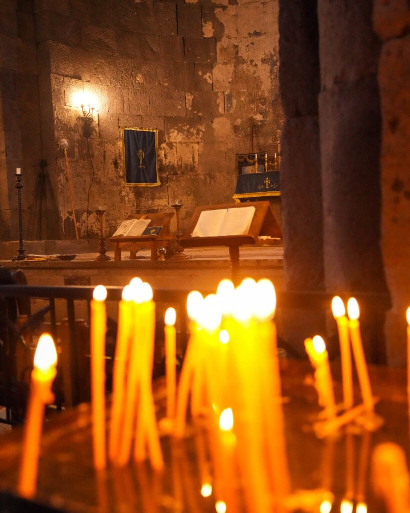 Im Vordergrund brennende Kerzen, ein Altar, Bücher und Banner in einer schummrigen Kirche mit Steinwänden.