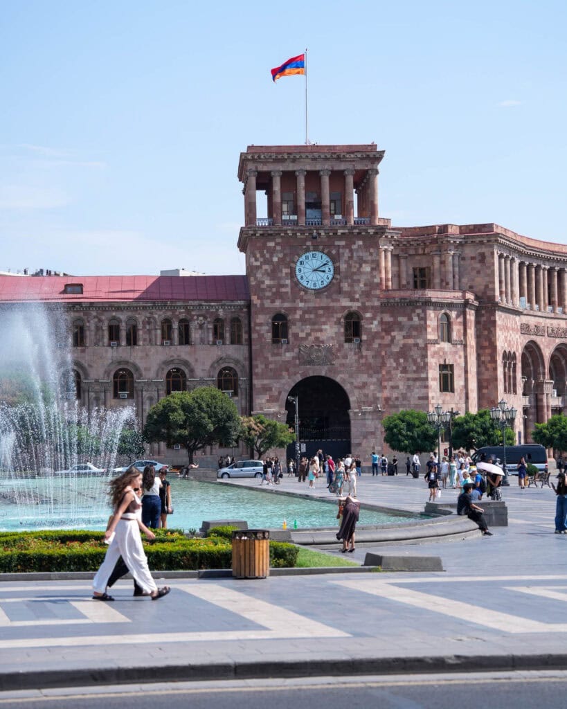 Menschen spazieren an einem großen Springbrunnen vorbei, im Hintergrund sind ein Uhrturm und die armenische Flagge zu sehen.