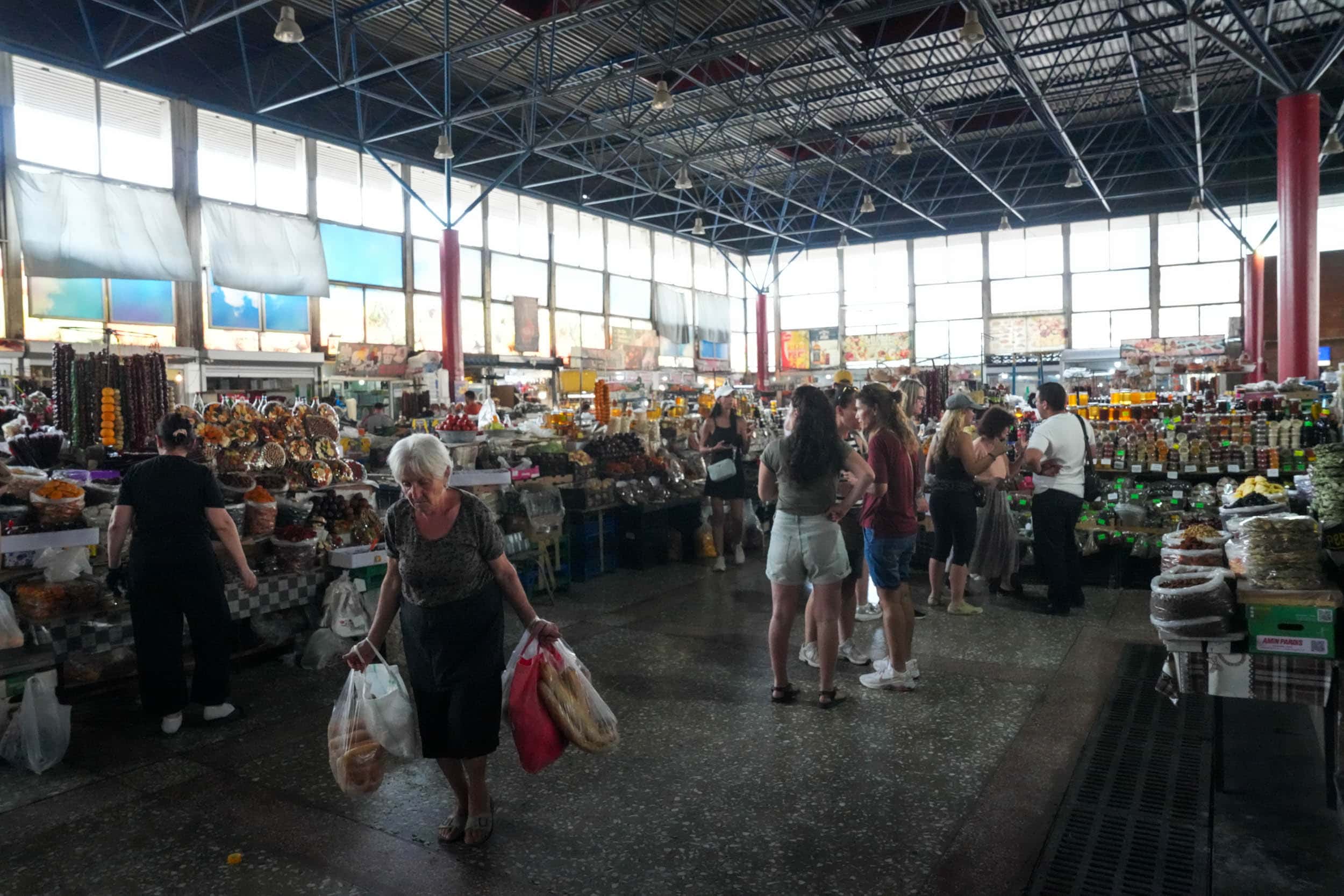 Menschen, die auf einem überdachten Markt, in den durch große Fenster Tageslicht fällt, Lebensmittel und Waren einkaufen.