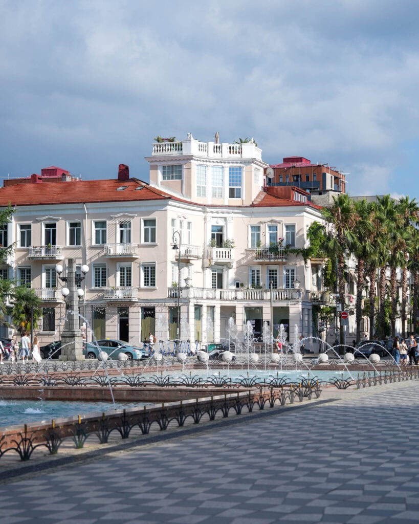 Stadtplatz in Batumi mit Springbrunnen, Palmen und hellen Gebäuden unter einem teilweise bewölkten Himmel.