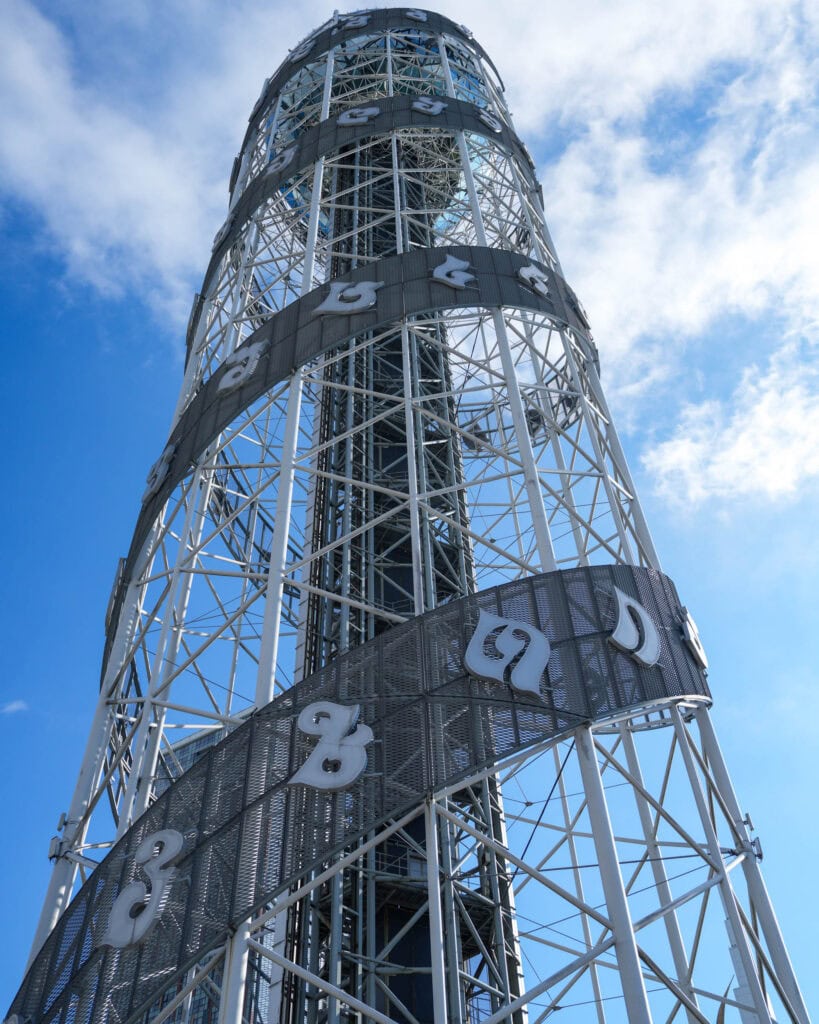 Ein hoher Metallturm mit spiralförmiger Struktur und Zahlen, der sich vor einem strahlend blauen Himmel mit vereinzelten Wolken abhebt, ist ein Wahrzeichen von Batumi.