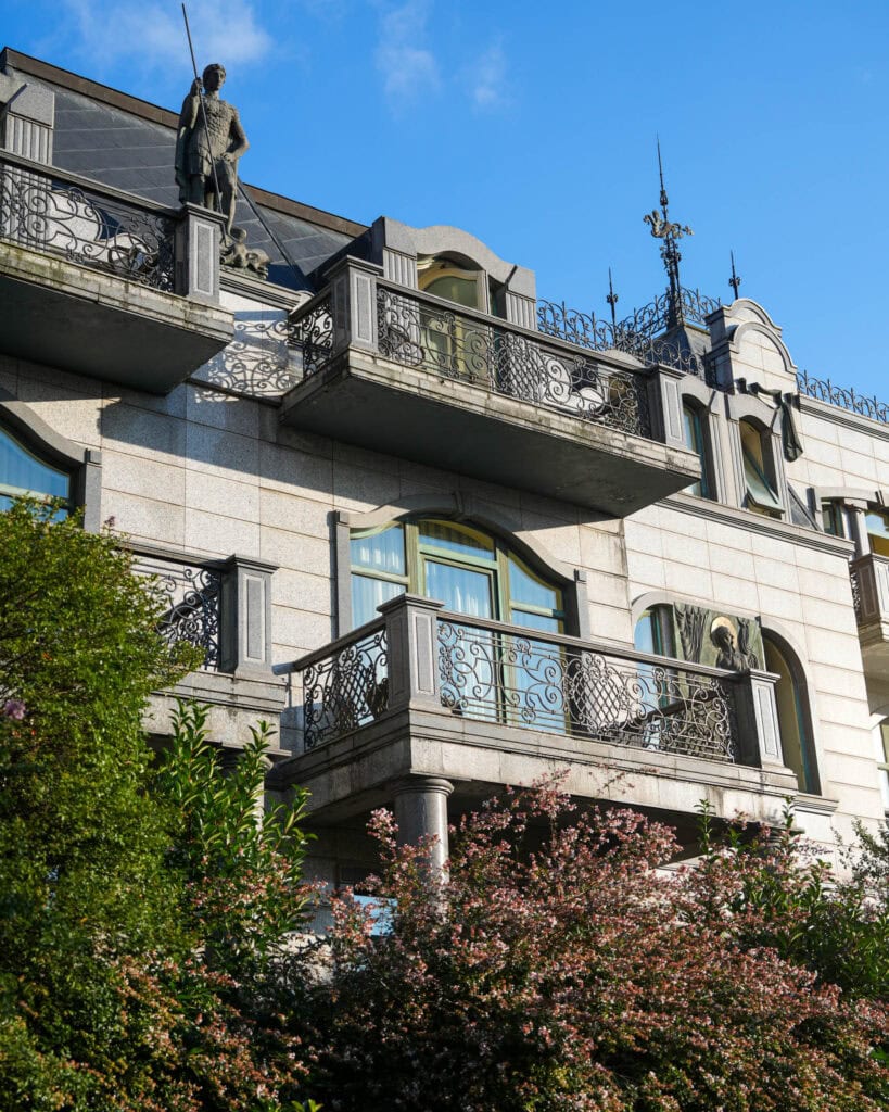 Ein prunkvolles Steingebäude mit Statuen auf den Balkonen, schmiedeeisernen Geländern und blühenden Sträuchern darunter, das an den eleganten architektonischen Charme Batumis erinnert.