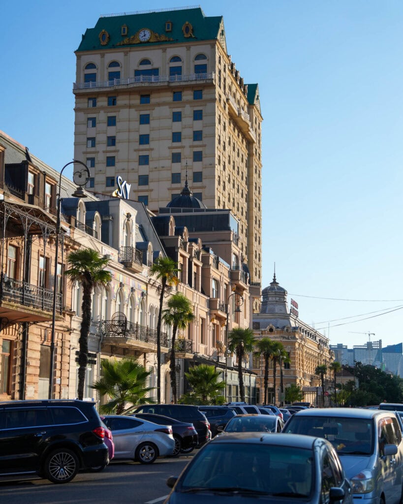 Autos fahren auf einer Straße in Batumi, die von Palmen und hohen historischen Gebäuden gesäumt ist, unter einem strahlend blauen Himmel.