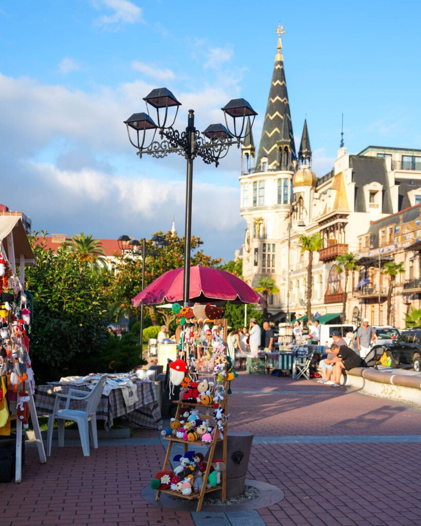 Straßenstand mit Stofftieren unter einem rosa Sonnenschirm in Batumi, im Hintergrund historische Gebäude und blauer Himmel.