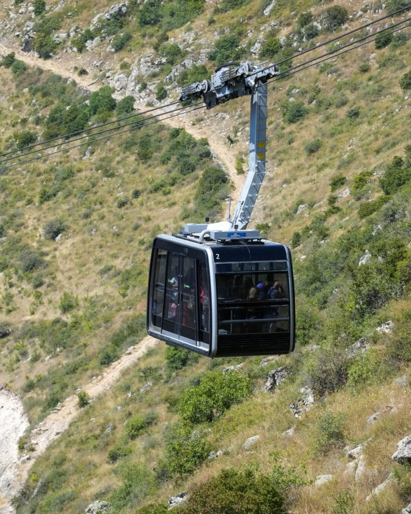 An einem sonnigen Tag fährt eine Seilbahn mit Passagieren an einem grasbewachsenen, felsigen Hang hinauf.