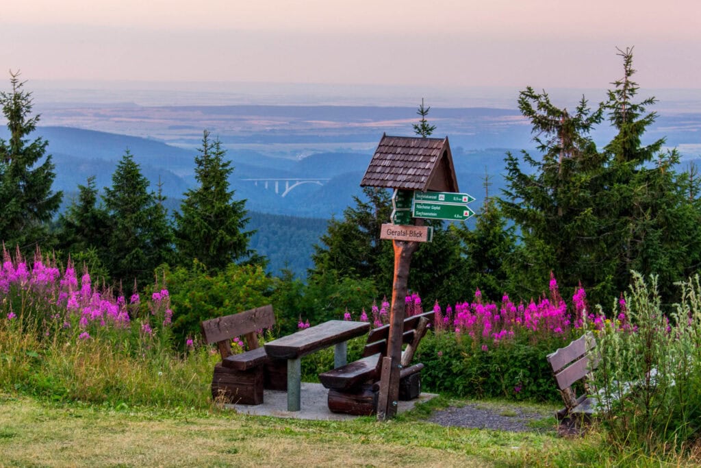 Holzbänke und ein Tisch mit einem Wegweiser, umgeben von Blumen, mit Blick auf Hügel und eine Brücke in der Ferne.