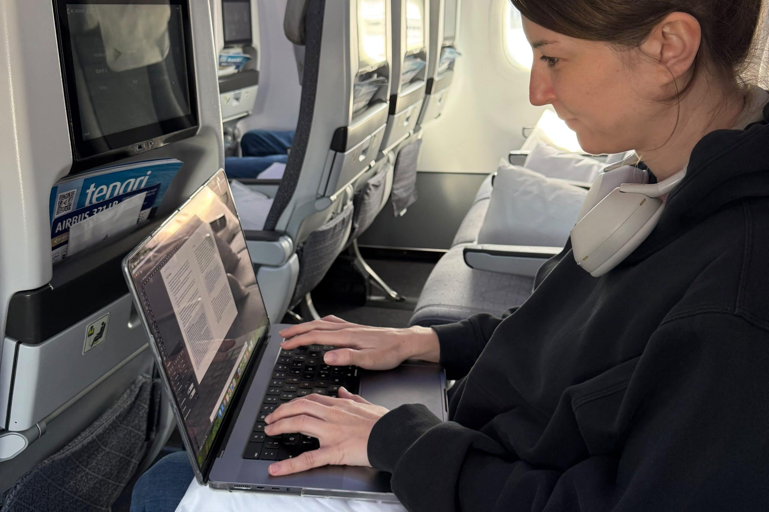 Eine Person, die auf einem Laptop einen Flugratgeber schreibt, während sie in einem Flugzeugsitz sitzt und die Kopfhörer um den Hals hängt.