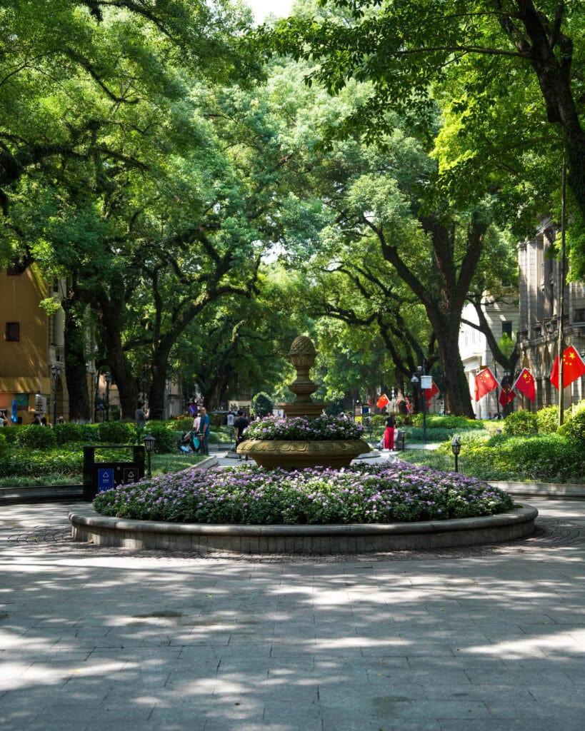 Ein rundes Beet, das einen Steinbrunnen in einem von Bäumen gesäumten Park mit chinesischen Flaggen umgibt.