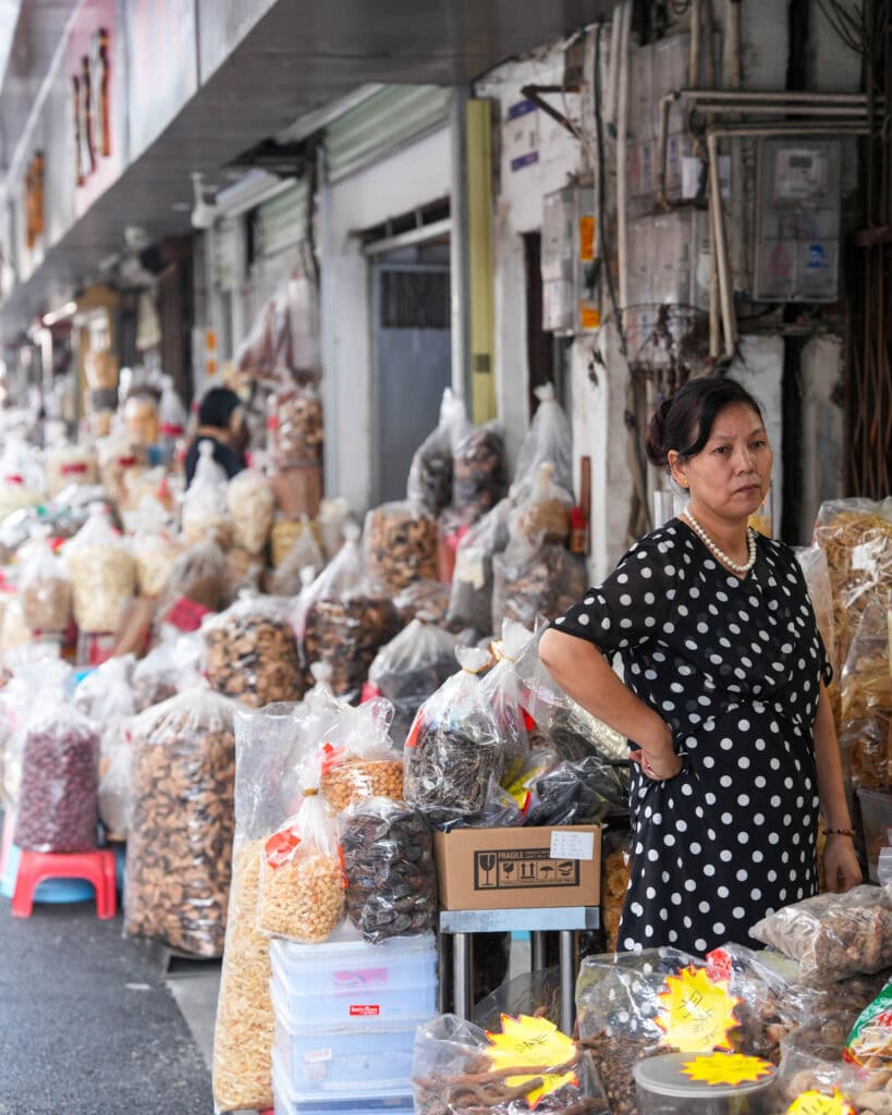 Eine Frau in einem schwarzen Kleid mit Punkten steht an einem Marktstand, an dem Tüten mit Trockenwaren verkauft werden.