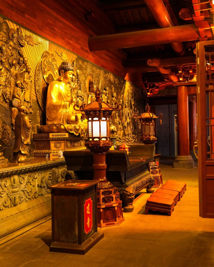 Eine goldene Buddha-Statue vor einer mit Schnitzereien verzierten Wand in einem schwach beleuchteten Tempel mit Holzlaternen.