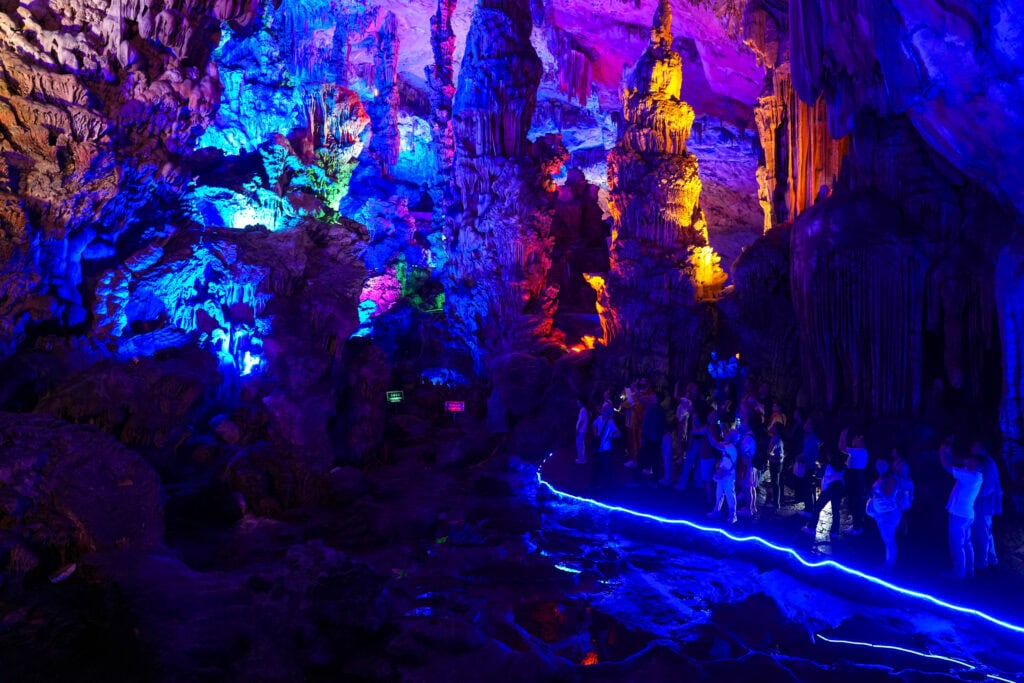 Menschen, die eine Kalksteinhöhle besichtigen, die von blauem, rosa und gelbem Licht beleuchtet wird.